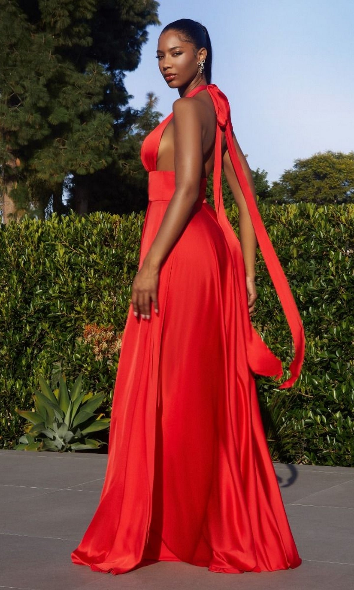A woman stands outdoors in greenery wearing the Long Infinity Prom Dress with Slit: Ladivine CD323, a bright red, floor-length A-line gown with a dramatic ribbon detail.